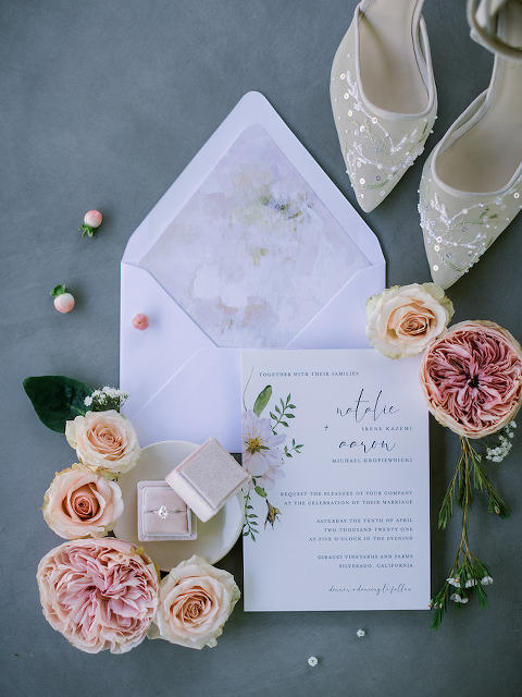Wedding invitation suite laid flat surrounded by fresh blush florals and a wedding ring and bridal shoes.