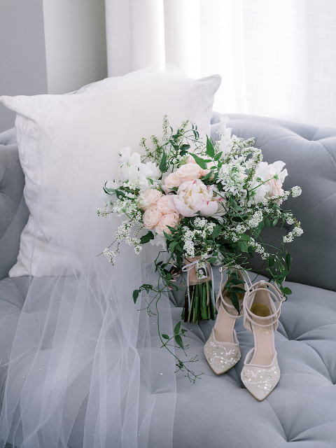Bridal bouquet, veil, and bridal shoes sitting on a tufted grey couch. 